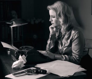 Black and white photograph of a blond woman sitting at a desk in front of a typewriter. The typewriter is surrounded by loose piles of paper, with a revolver sitting on top of one pile.